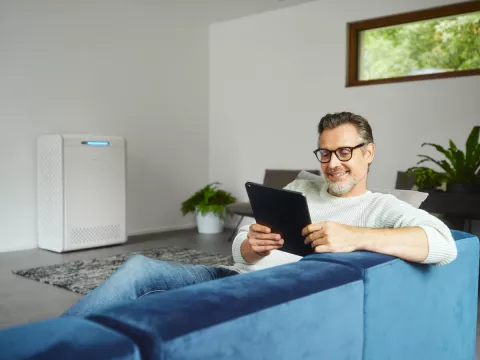 Ein Mann sitzt auf einem Sofa im Wohnzimmer, während im Hintergrund sein neu gekaufter Stromspeicher steht.